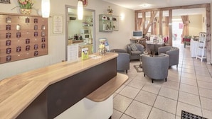 A reception area with a wooden counter, grey armchairs, a small table with a TV, and a kitchen in the background. - Hôtel Restaurant le Pan de Bois (Breviandes)