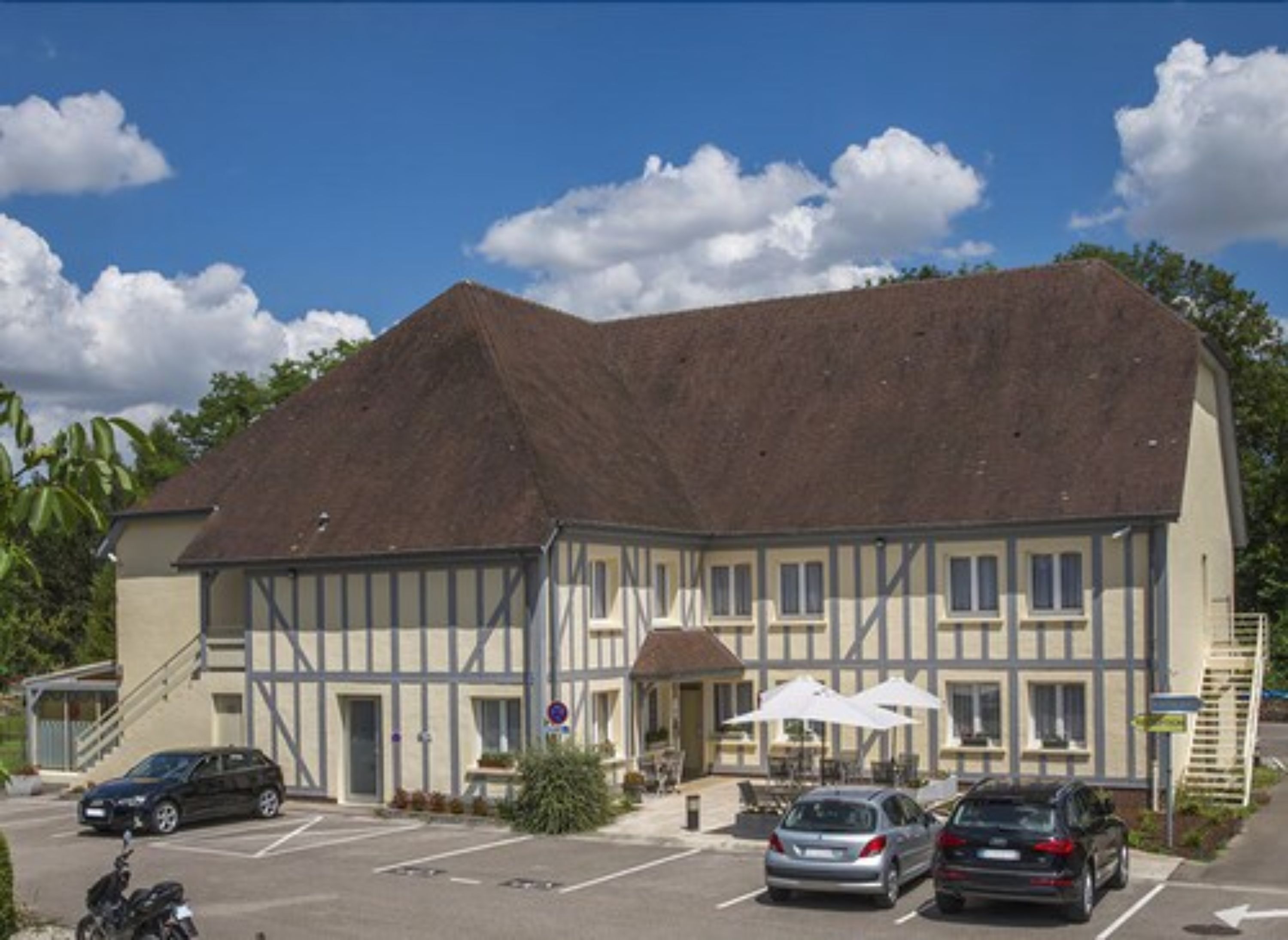 Ein Gebäude mit einem steilen Dach, einem Parkplatz mit mehreren Autos und Sitzgelegenheiten im Freien mit Sonnenschirmen.