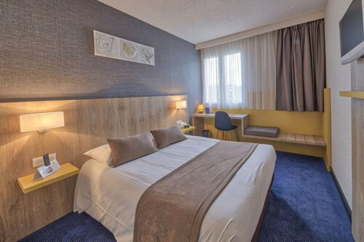 A hotel room with a bed, bedside table, lamp, desk, chair, and a window with curtains.