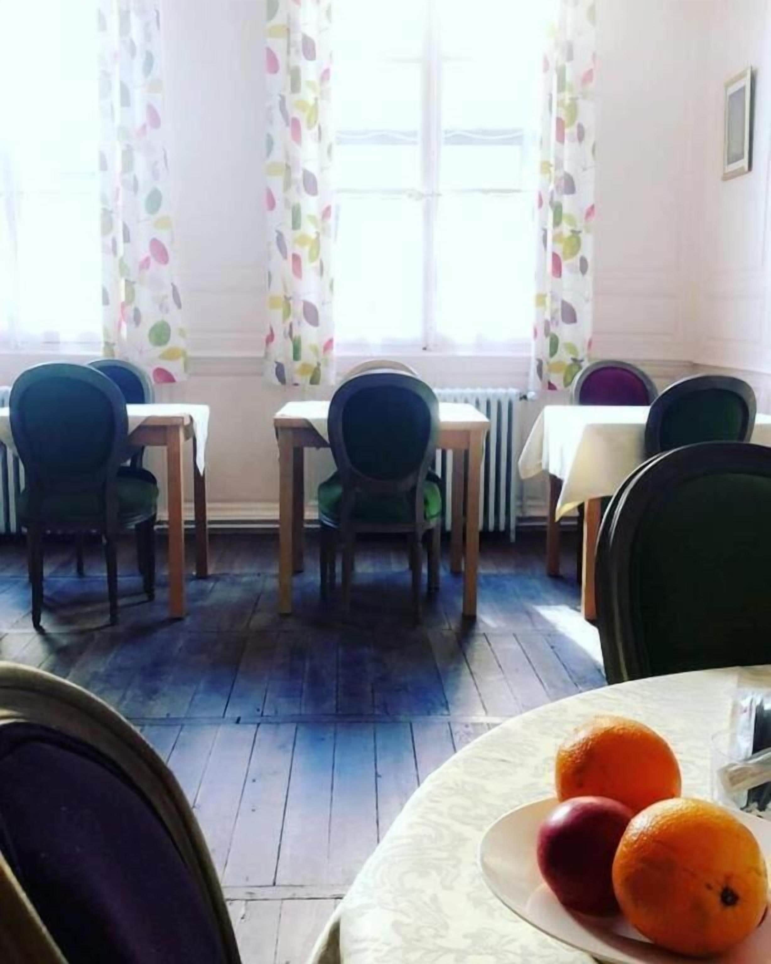 Un coin repas avec des tables et des chaises, une fenêtre avec des rideaux et une assiette de fruits posée sur une table.