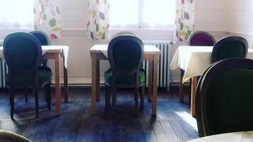 Un coin repas avec des tables et des chaises, une fenêtre avec des rideaux et une assiette de fruits posée sur une table.