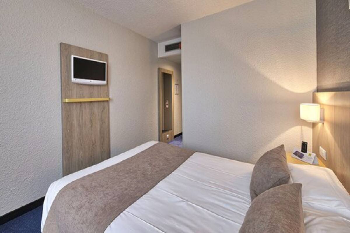 A hotel room with a bed, a TV mounted on the wall, a bedside lamp, and a door leading to another room.