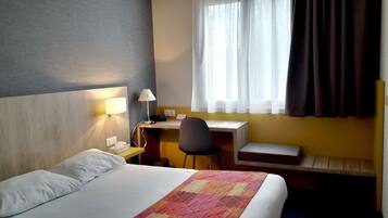 A hotel room with a bed, a desk, a chair, a lamp, and a window with curtains.
