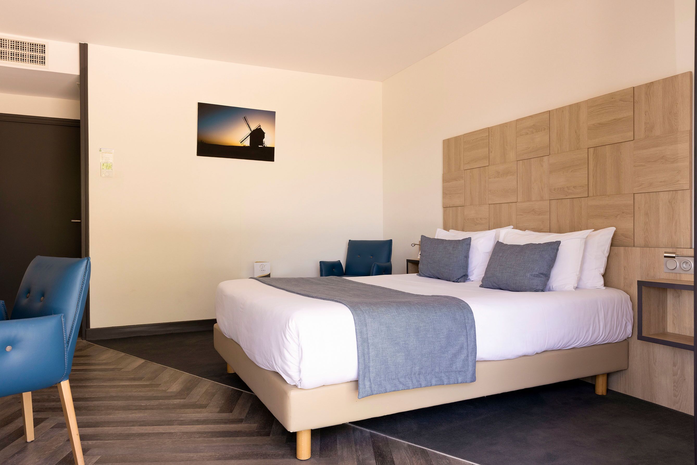 Ein modernes Hotelzimmer mit einem großen Bett, einem blauen Sessel, einem hölzernen Kopfteil und einem Bild von einem Windrad an der Wand.