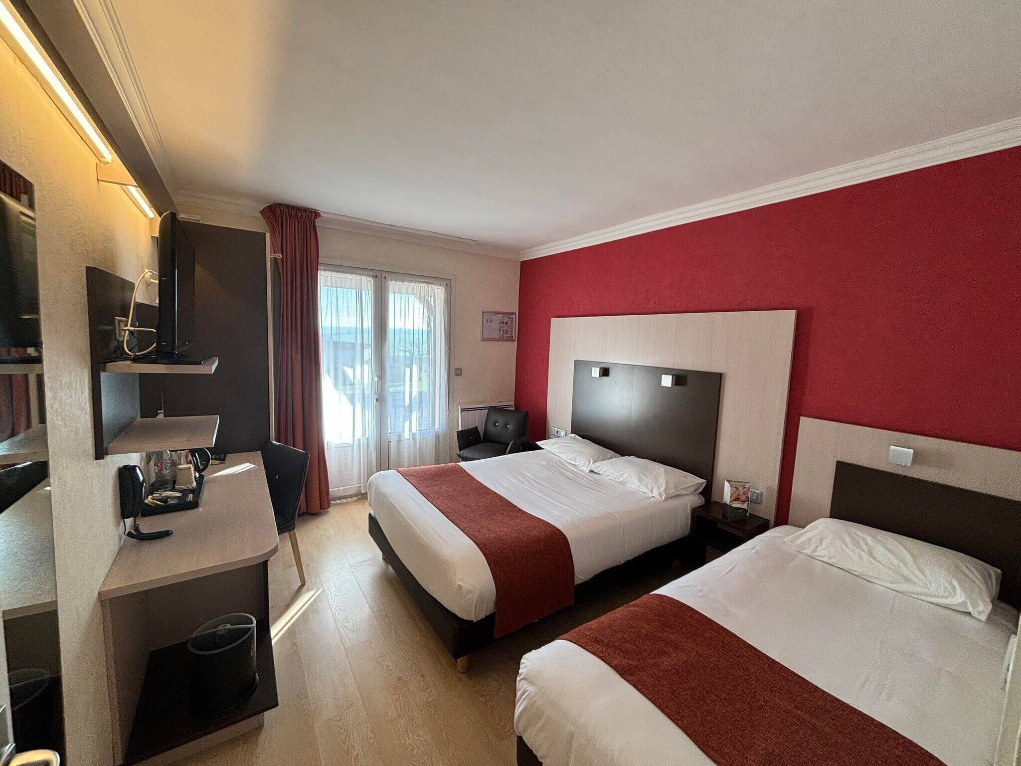 Une chambre d’hôtel avec deux lits, un bureau avec une chaise, une télévision et une fenêtre avec des rideaux.
