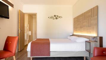 A hotel room with a bed, two red chairs, a wooden headboard, a wall-mounted TV, and a decorative wall piece.