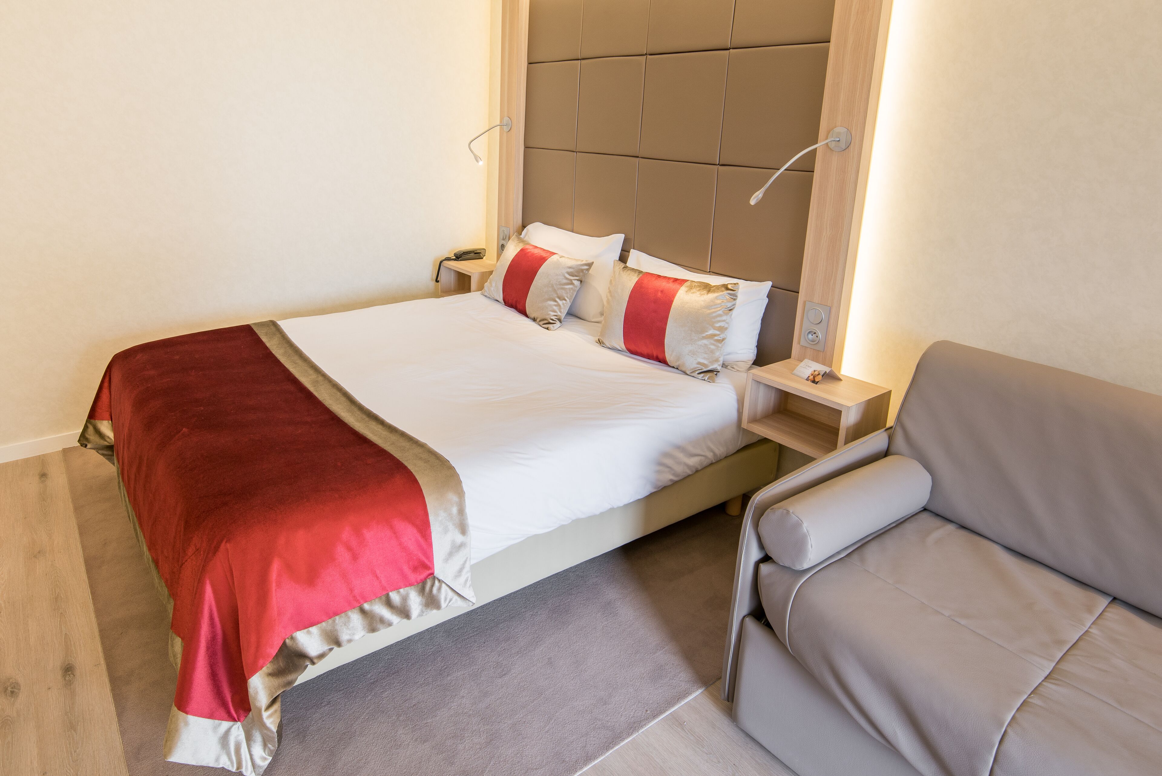 A modern hotel room with a large bed, a grey sofa, and a wooden floor.