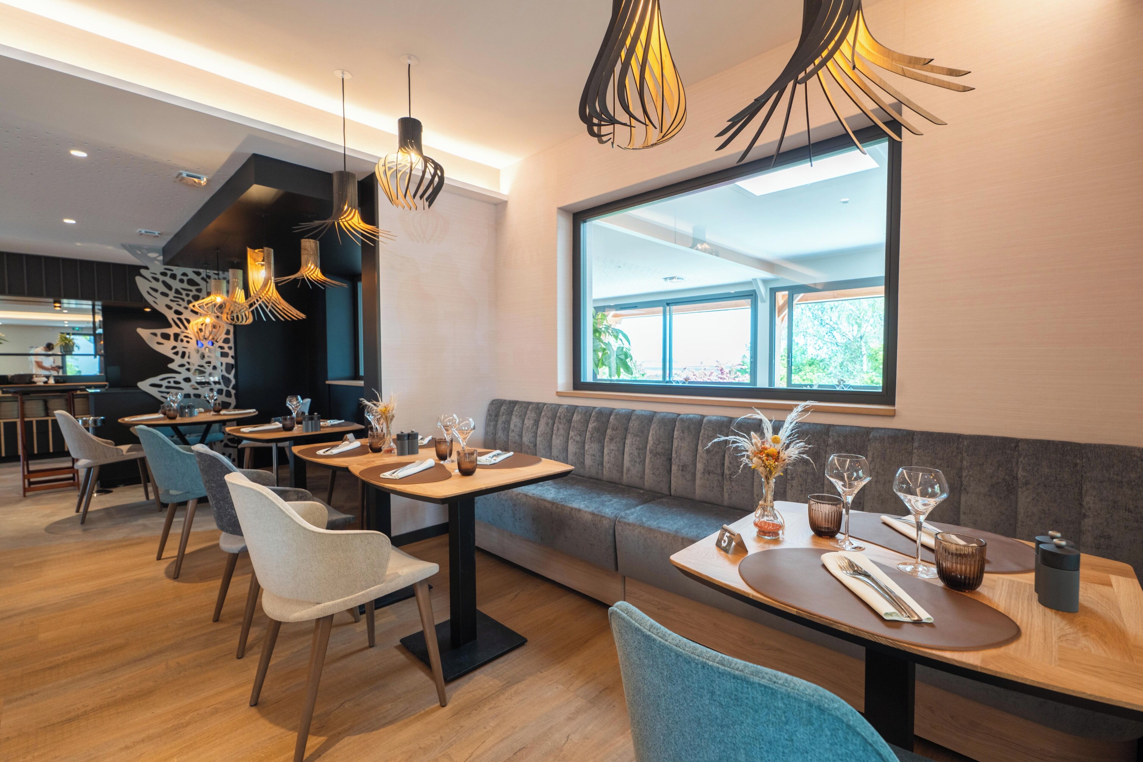 Ein modernes Restaurant mit einem langen Esstisch, gepolsterten Sitzplätzen, Pendelleuchten und einem großen Fenster.