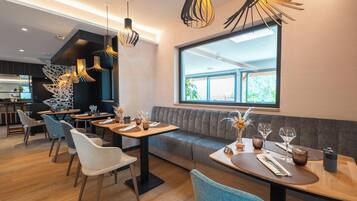 Ein modernes Restaurant mit einem langen Esstisch, gepolsterten Sitzplätzen, Pendelleuchten und einem großen Fenster.