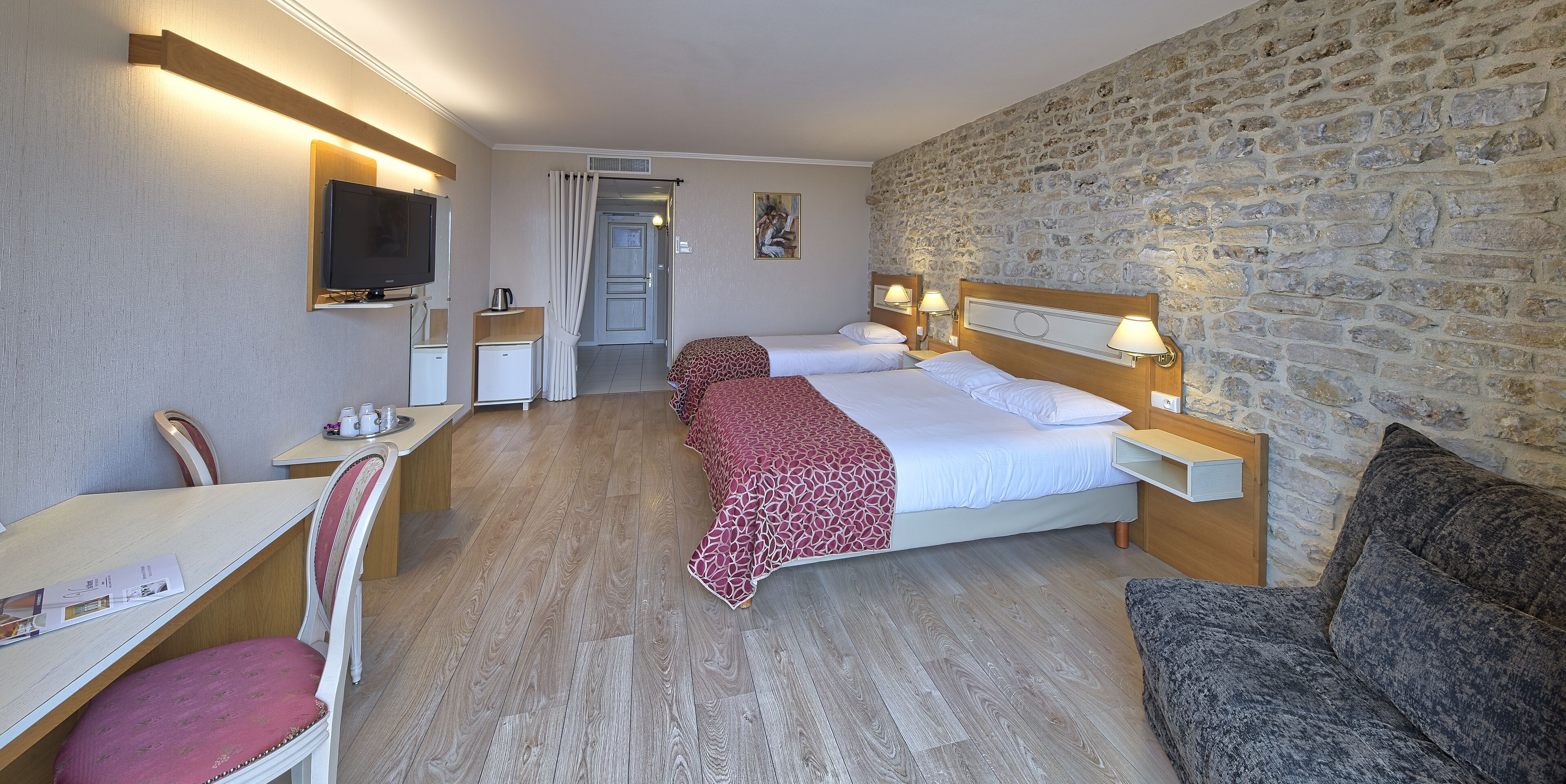 A hotel room with two beds, a desk with a chair, a television, and a stone wall.