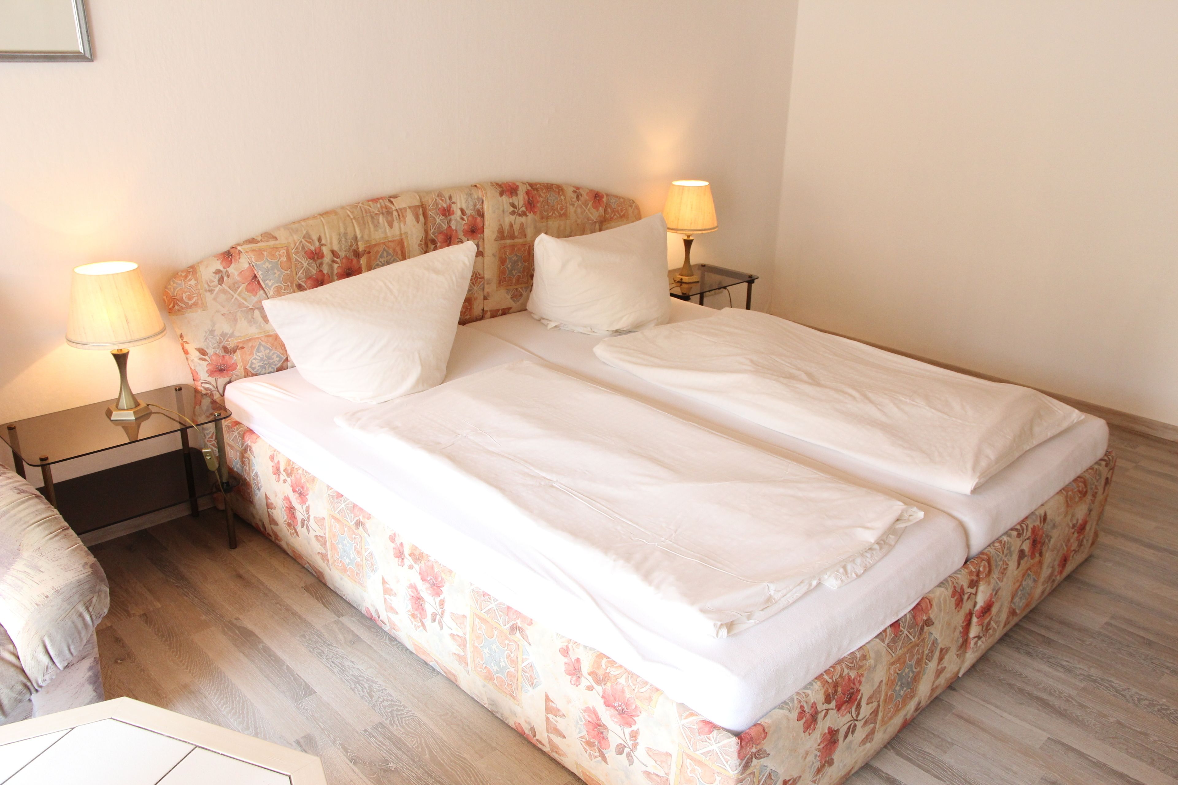 A neatly made bed with floral bedding, two bedside lamps, and a wooden floor.