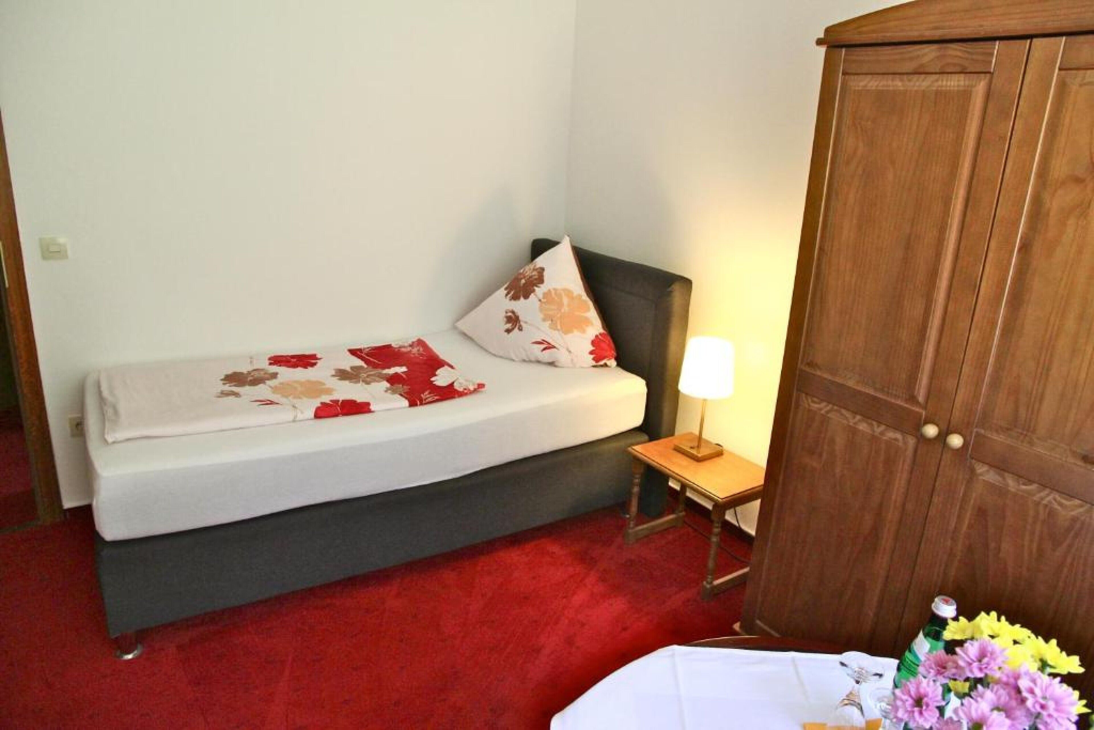 A bedroom with a bed, a nightstand with a lamp, a wooden wardrobe, and a red carpet.