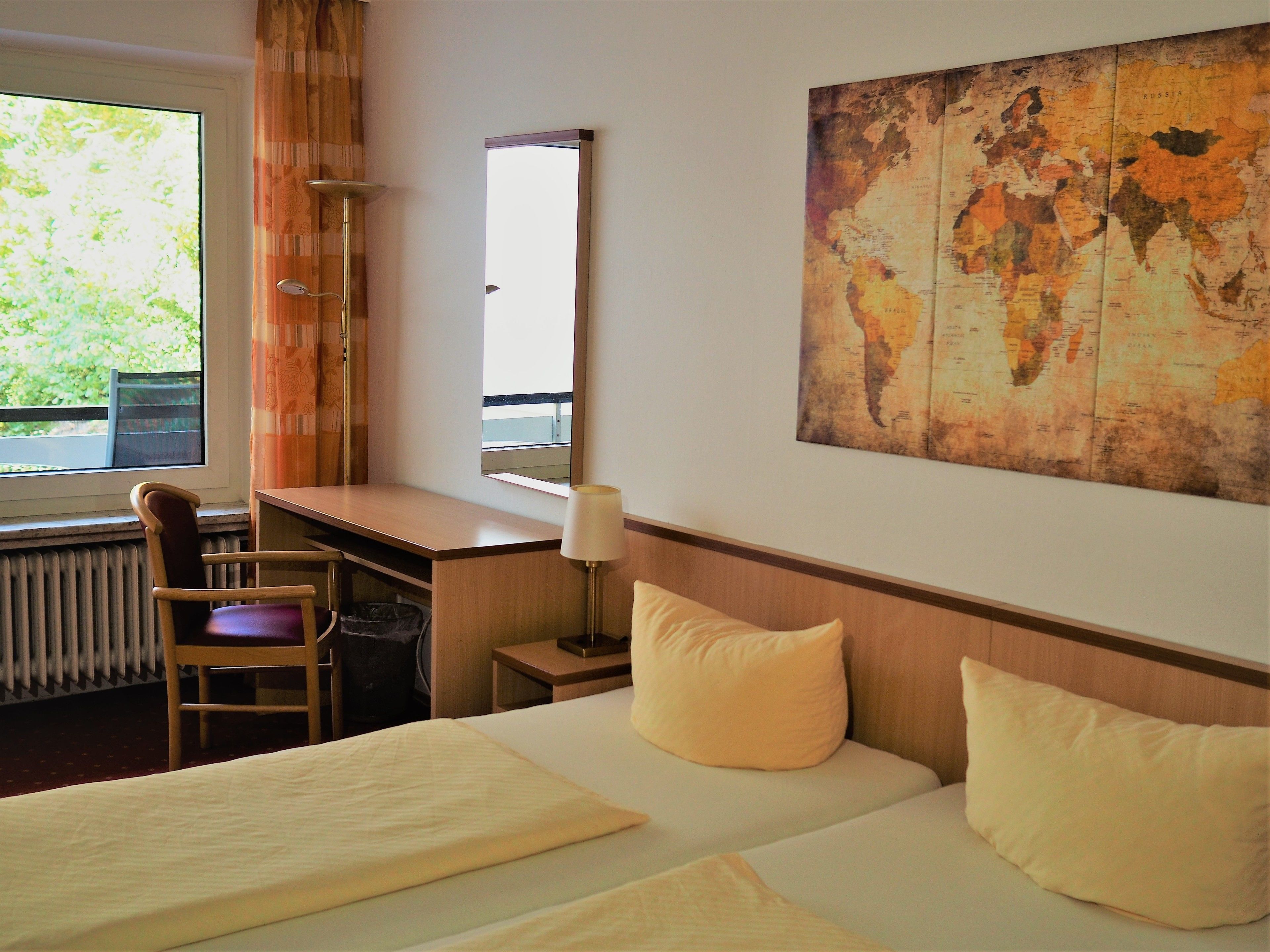 A hotel room with a bed, a desk, a chair, a lamp, and a large world map on the wall.