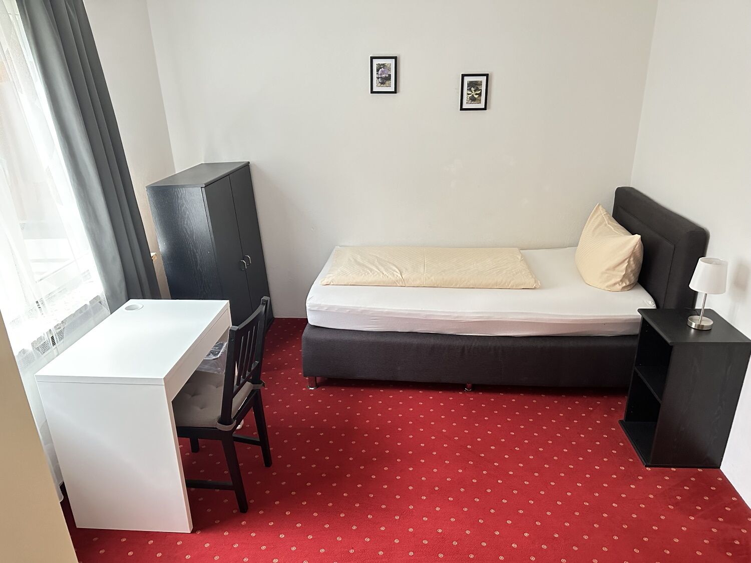 Ein kleines, ordentlich eingerichtetes Schlafzimmer mit Bett, Schreibtisch und Stuhl. An der Wand hängen zwei gerahmte Bilder.