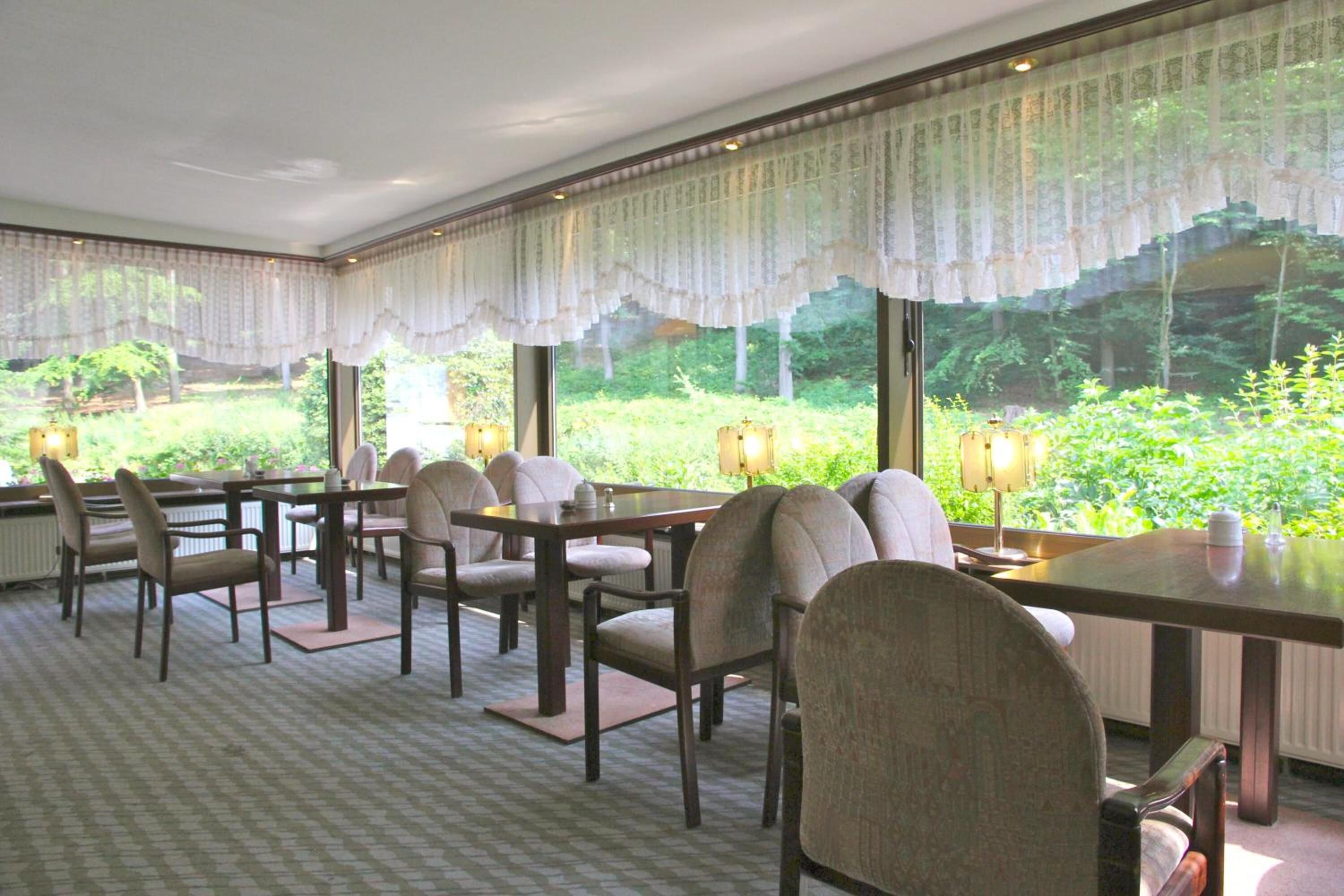 Un coin repas avec une table et des chaises, de grandes fenêtres ornées de rideaux en dentelle et une vue sur la verdure environnante.