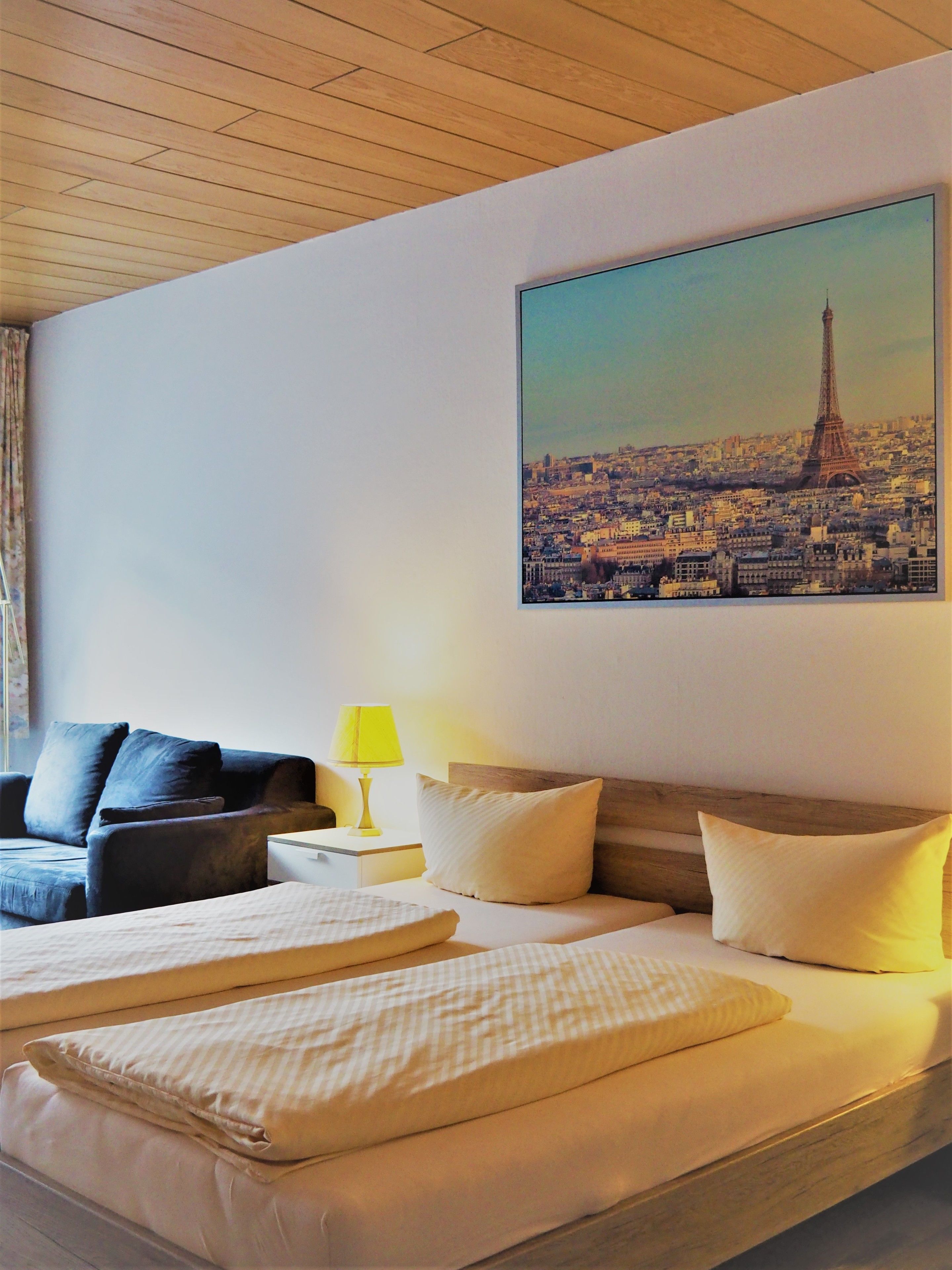 A hotel room with a bed, a couch, a nightstand with a lamp, and a cityscape painting on the wall.