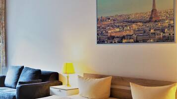 A hotel room with a bed, a couch, a nightstand with a lamp, and a cityscape painting on the wall.