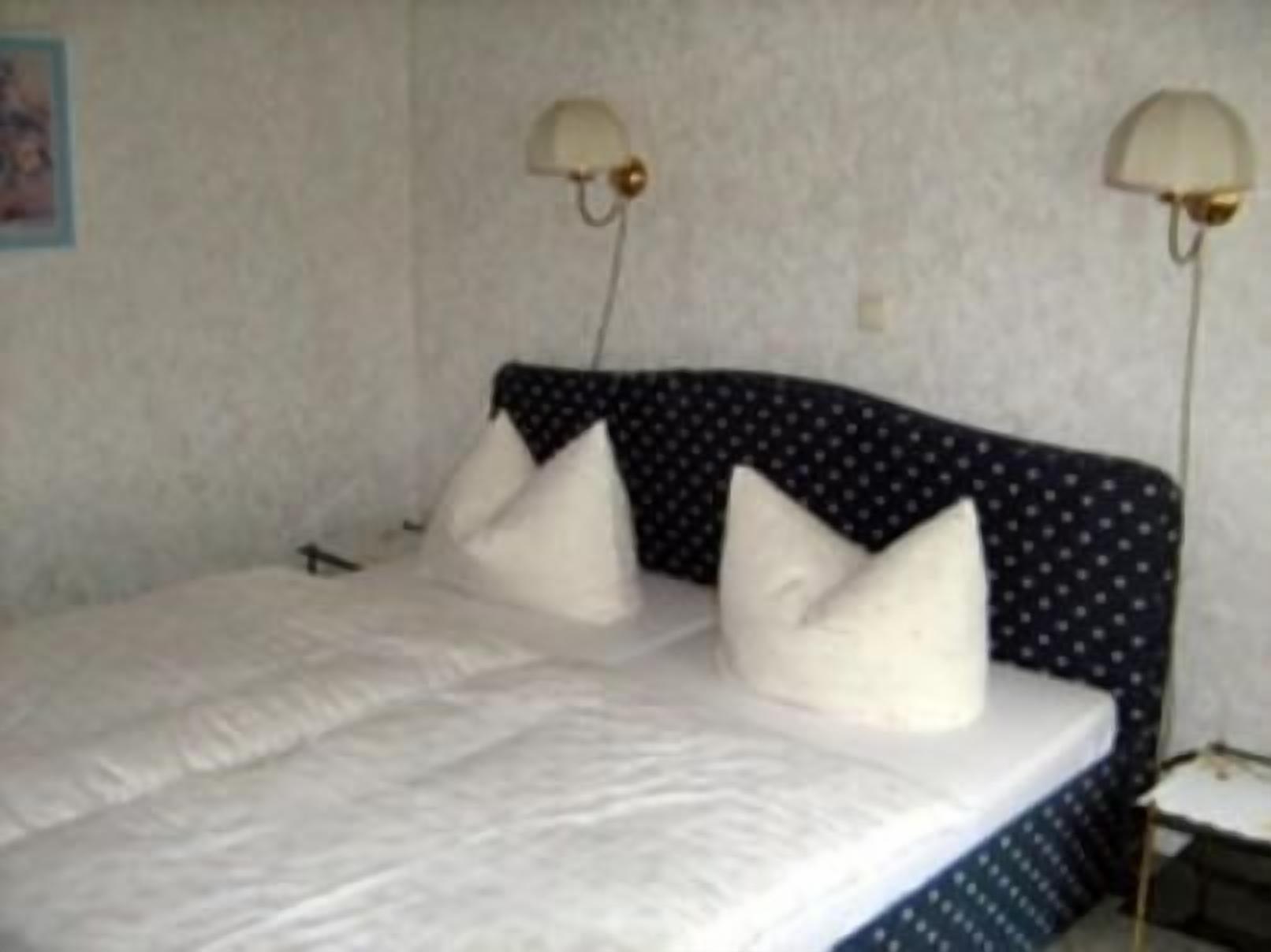 Una cama con edredón blanco y almohadas, cabecera oscura con estampado de lunares y dos lámparas fijadas a la pared.