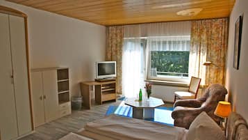 Una habitación con cama, televisión sobre un mueble bajo, una silla, una mesa con lámpara y una ventana con cortinas.