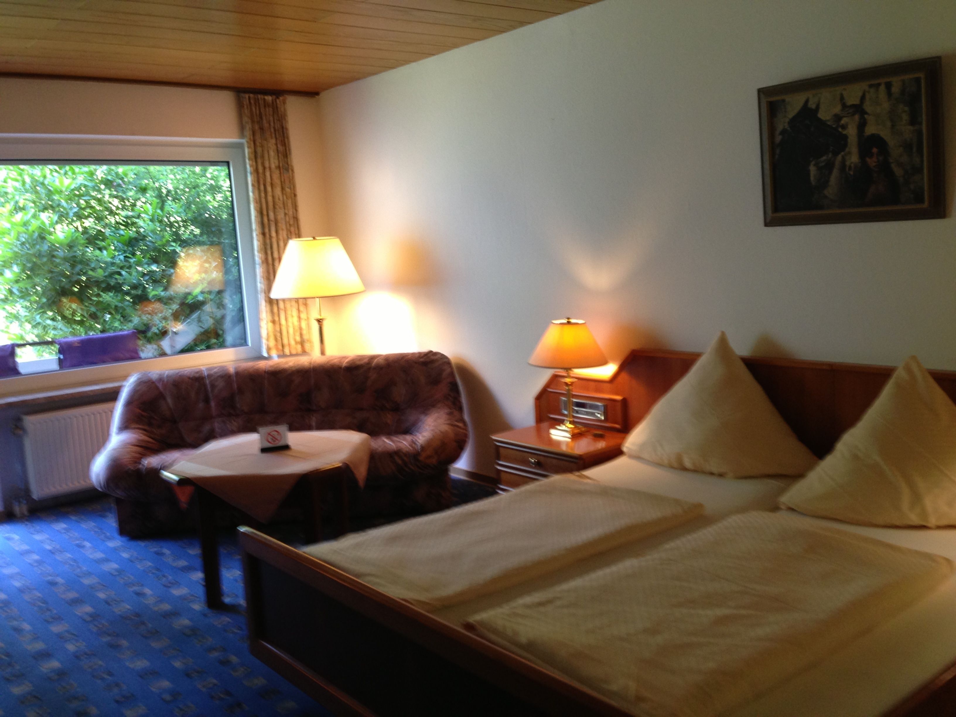 Een hotelkamer met een bed, een kleine tafel, een stoel, een lamp en een raam met uitzicht op groen.