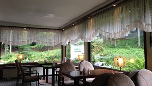A dining area with a table and chairs, large windows with lace curtains, and a view of a garden. - Waldhotel Porta Westfalica garni (Porta Westfalica)