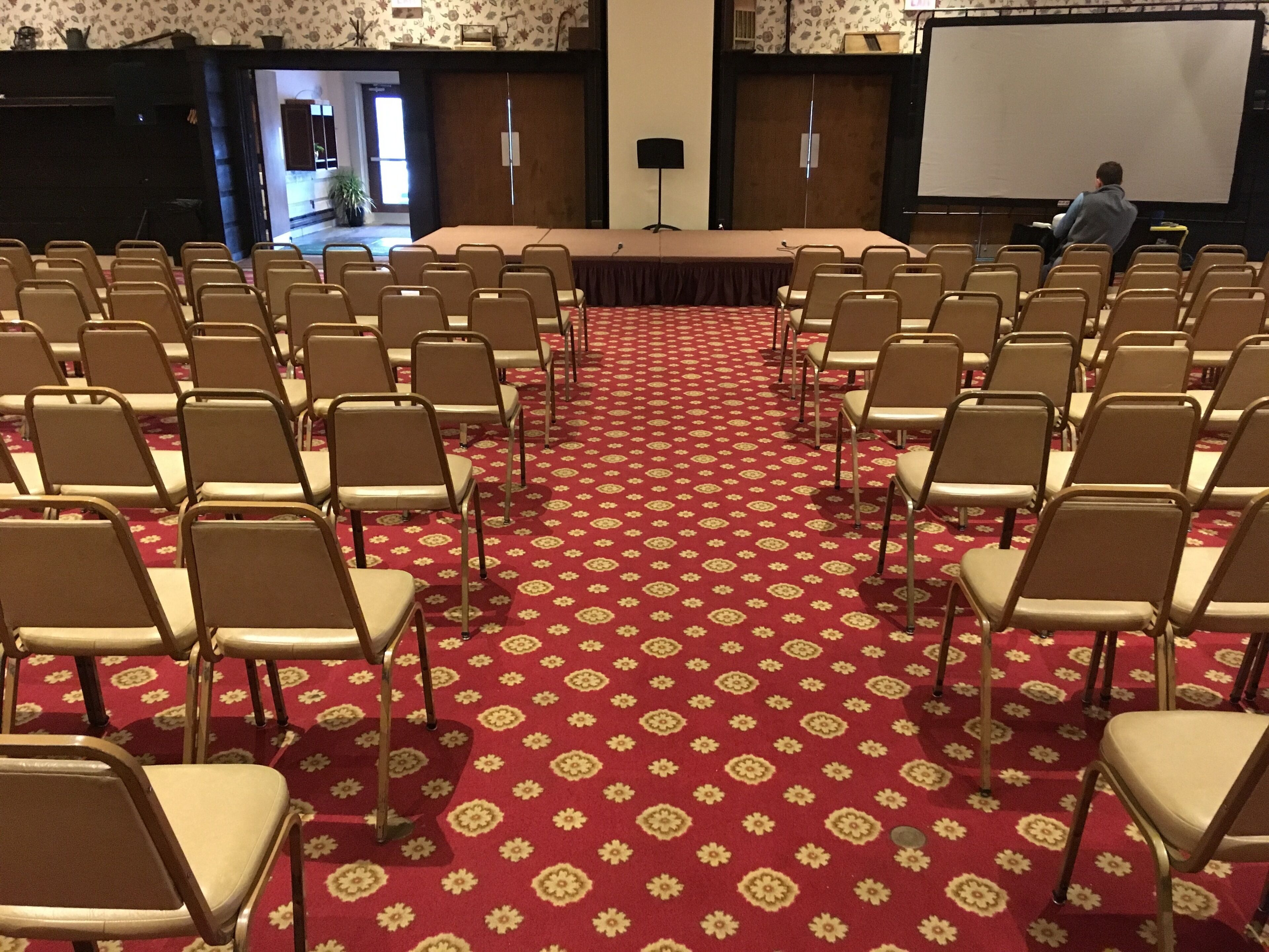 Un salón de conferencias con filas de sillas orientadas hacia una gran mesa y una pantalla de proyector.