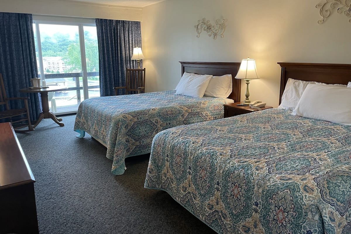 A hotel room with two beds, a desk, a chair, and a view of the outdoors through a large window.