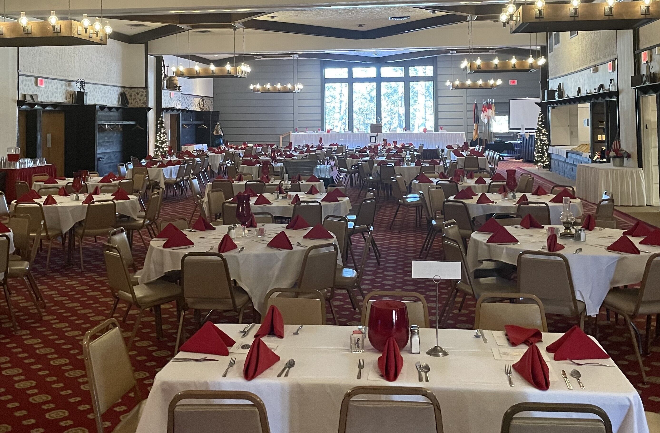 Uma sala de banquetes com várias mesas redondas dispostas para refeições, cada uma com toalhas brancas e guardanapos vermelhos.