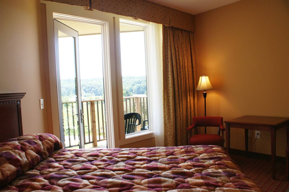 A hotel room with a large bed, a chair, a table, and a view of a balcony with a chair.