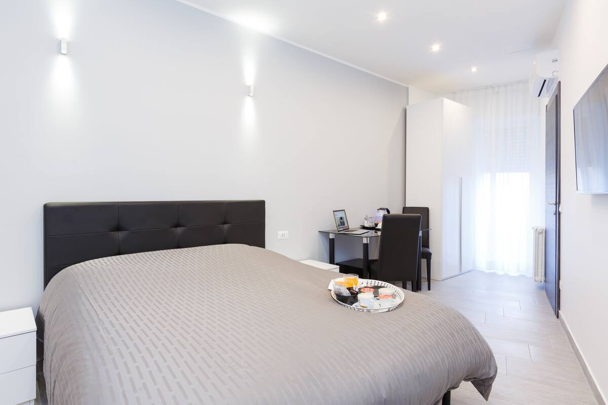 A modern hotel room with a large bed, a desk with a laptop, and a tray with breakfast items.