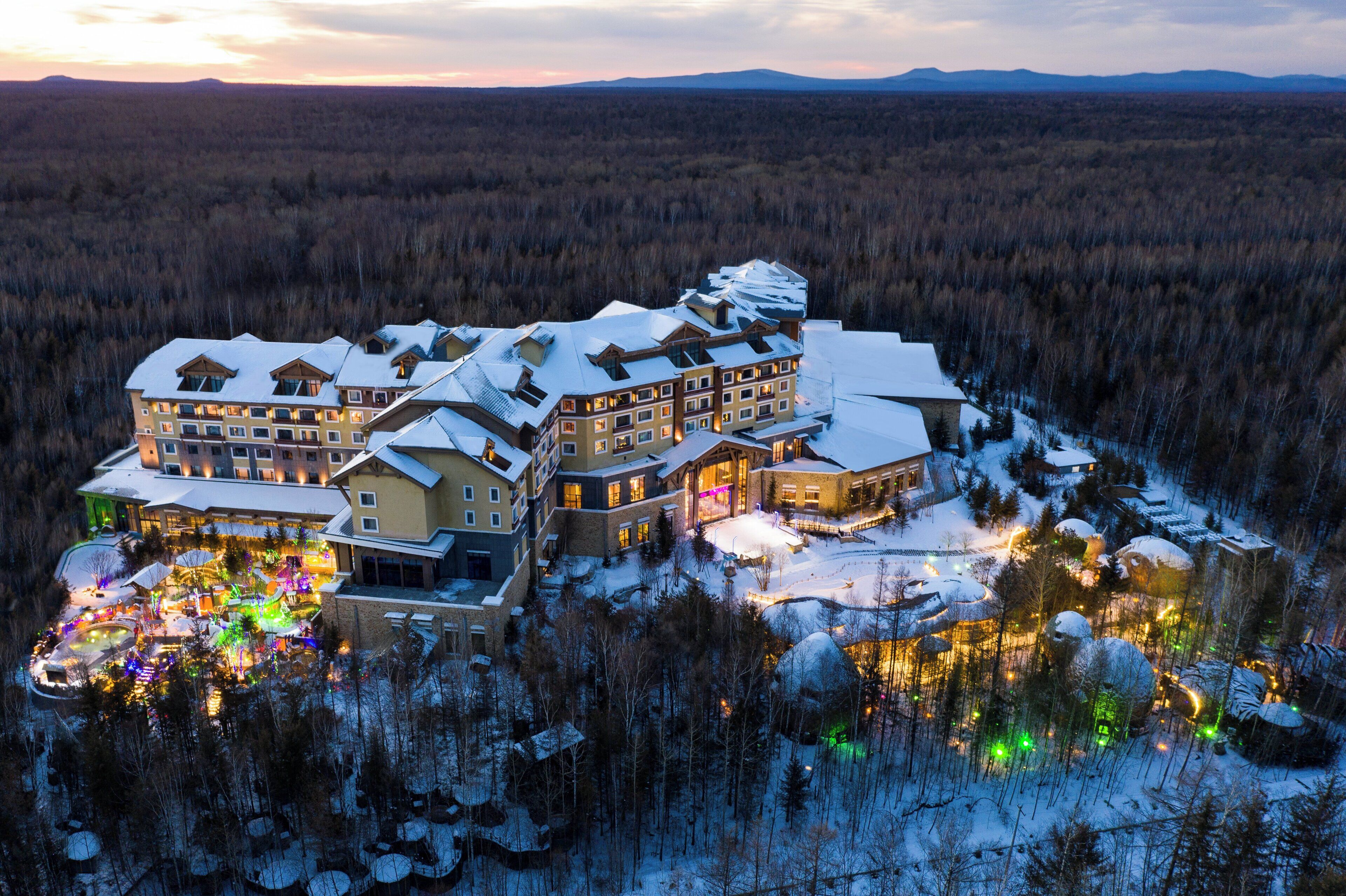 Um grande resort com vários edifícios, telhados cobertos de neve e um cenário de floresta.