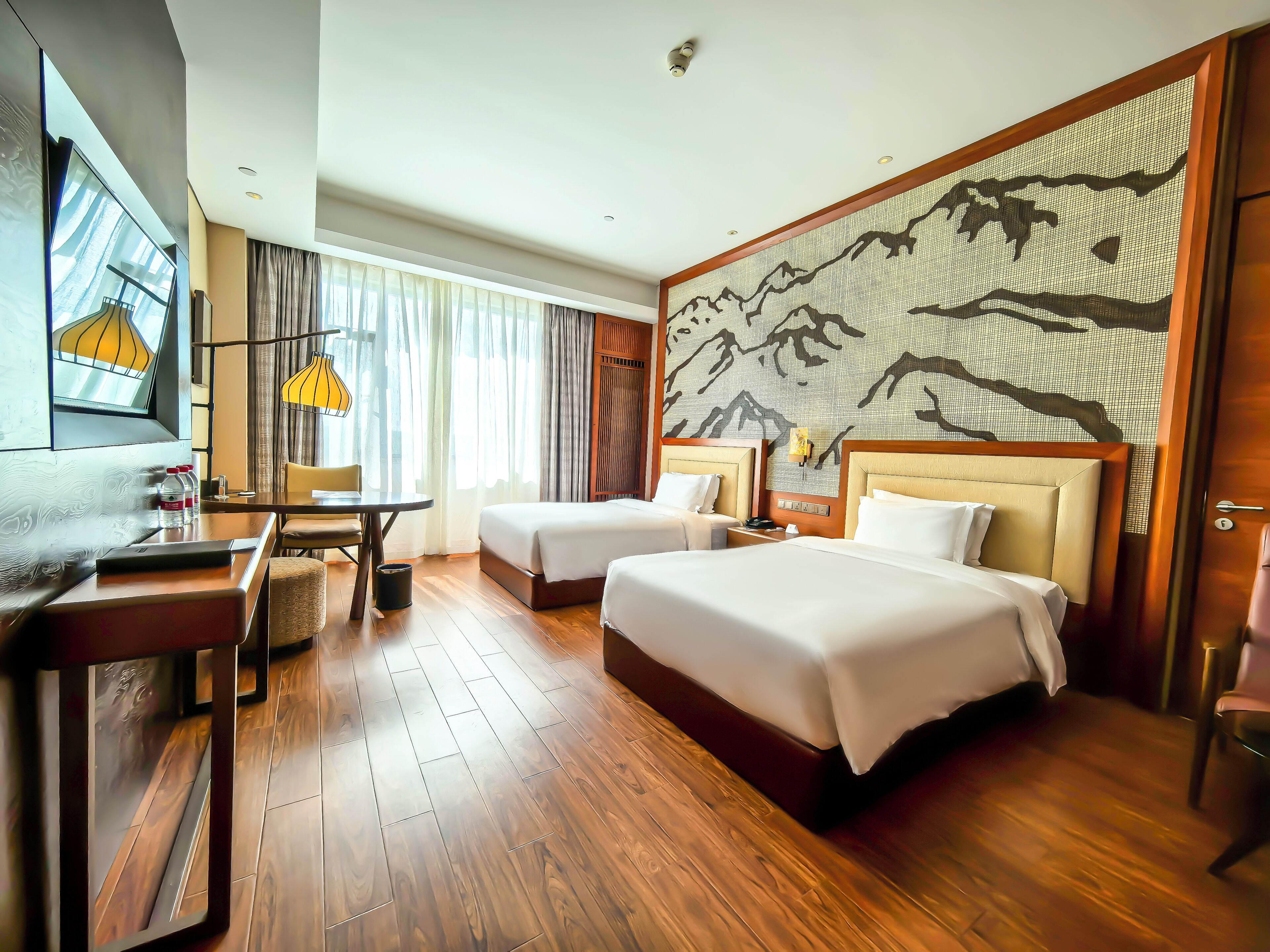 A hotel room with two beds, a desk, a chair, a table, and a large wall mural.