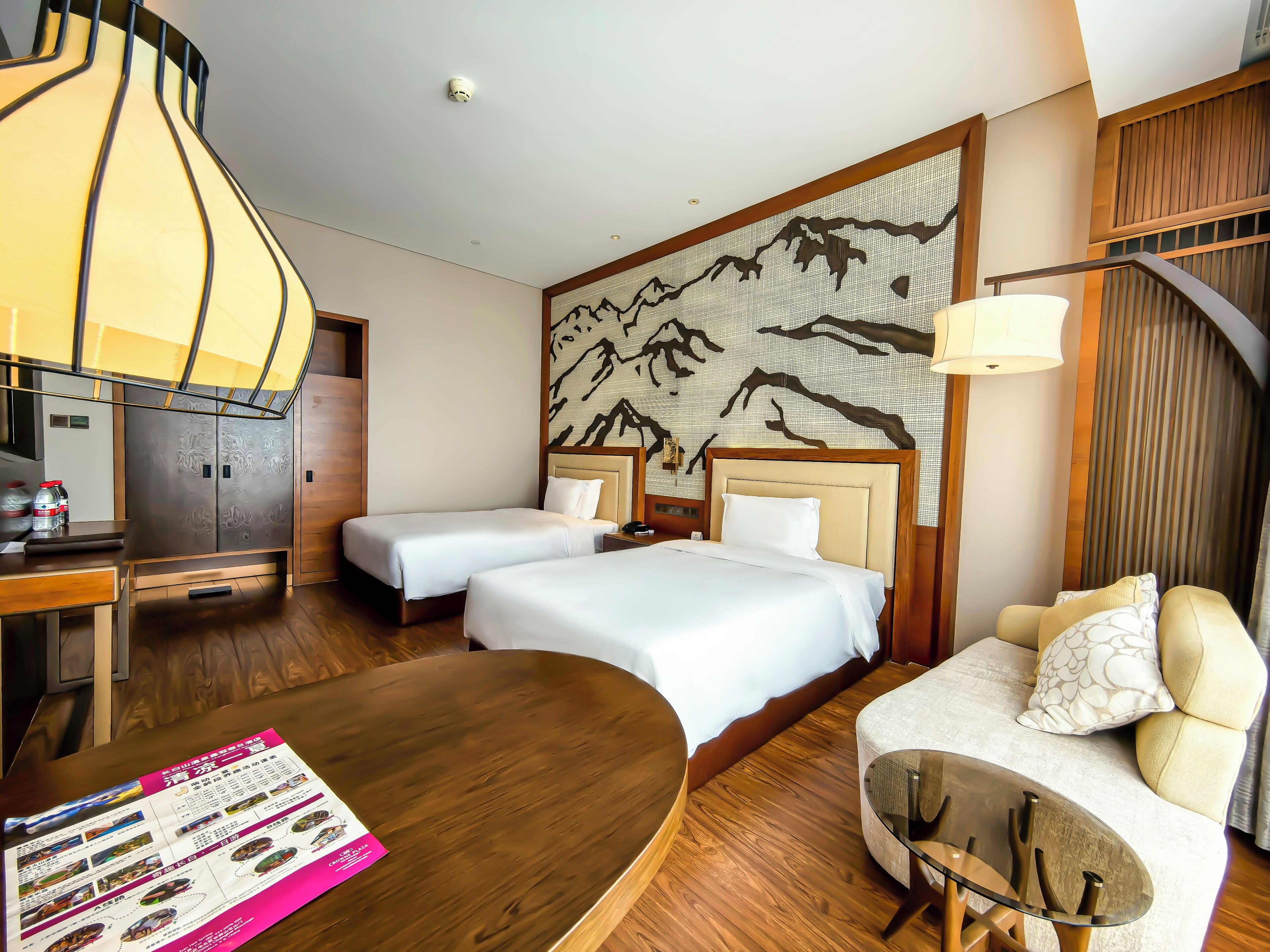 Quarto de hotel com duas camas, uma escrivaninha, um sofá, uma parede com desenho de montanha e um ventilador de teto.