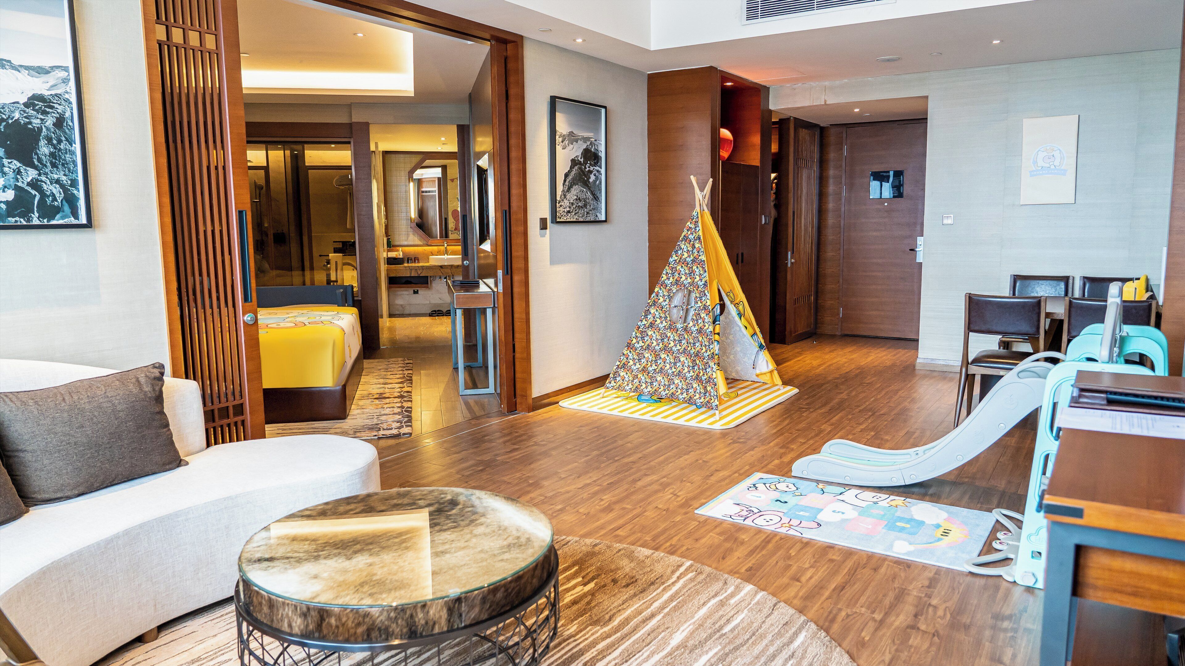 Una habitación de hotel moderna con sofá, una pequeña mesa redonda, una zona de juegos con tobogán y suelo de madera.