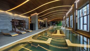 A modern indoor swimming pool with a curved design, lounge chairs, and a wooden ceiling with linear lighting.