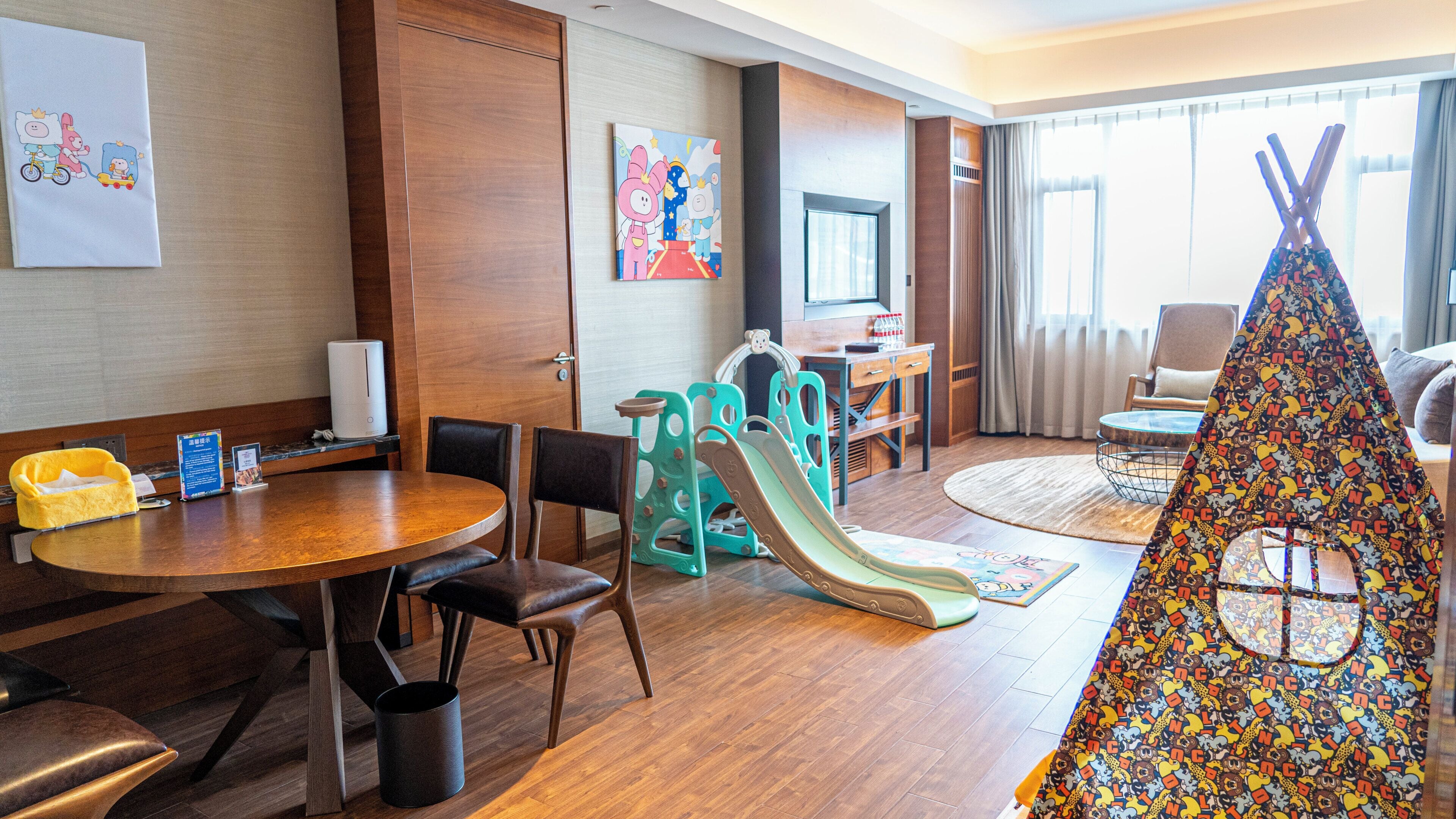 A hotel room with a wooden floor, a dining table with chairs, a children's play area with a slide, a sofa, a TV, and a window with curtains.