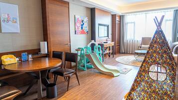 A hotel room with a wooden floor, a dining table with chairs, a children's play area with a slide, a sofa, a TV, and a window with curtains.