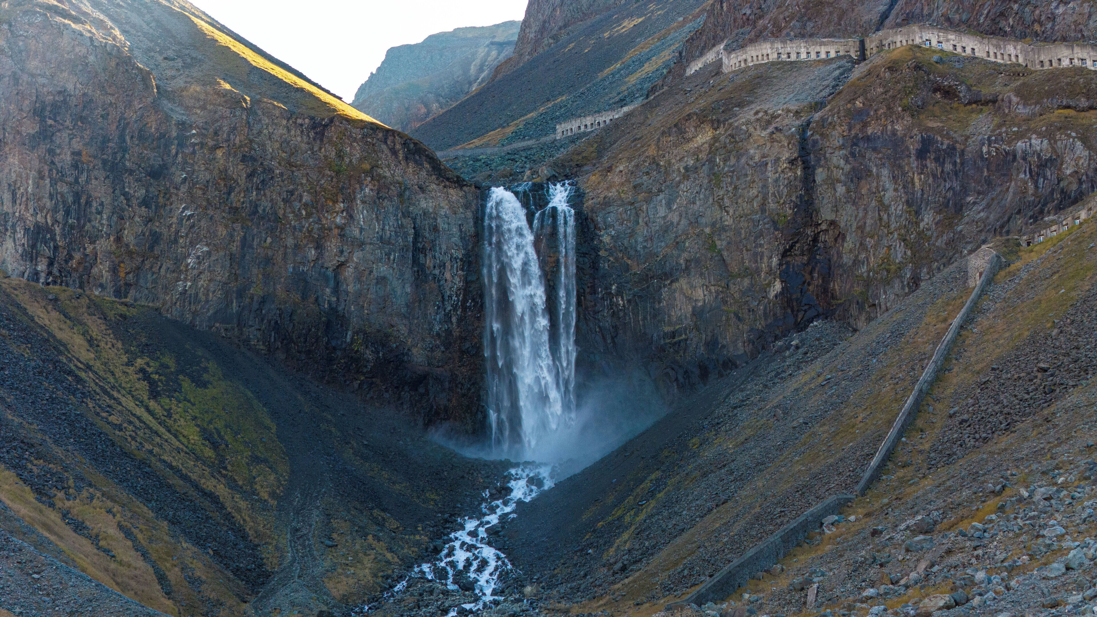 A waterfall cascading down a rocky valley with a stone structure on the right.