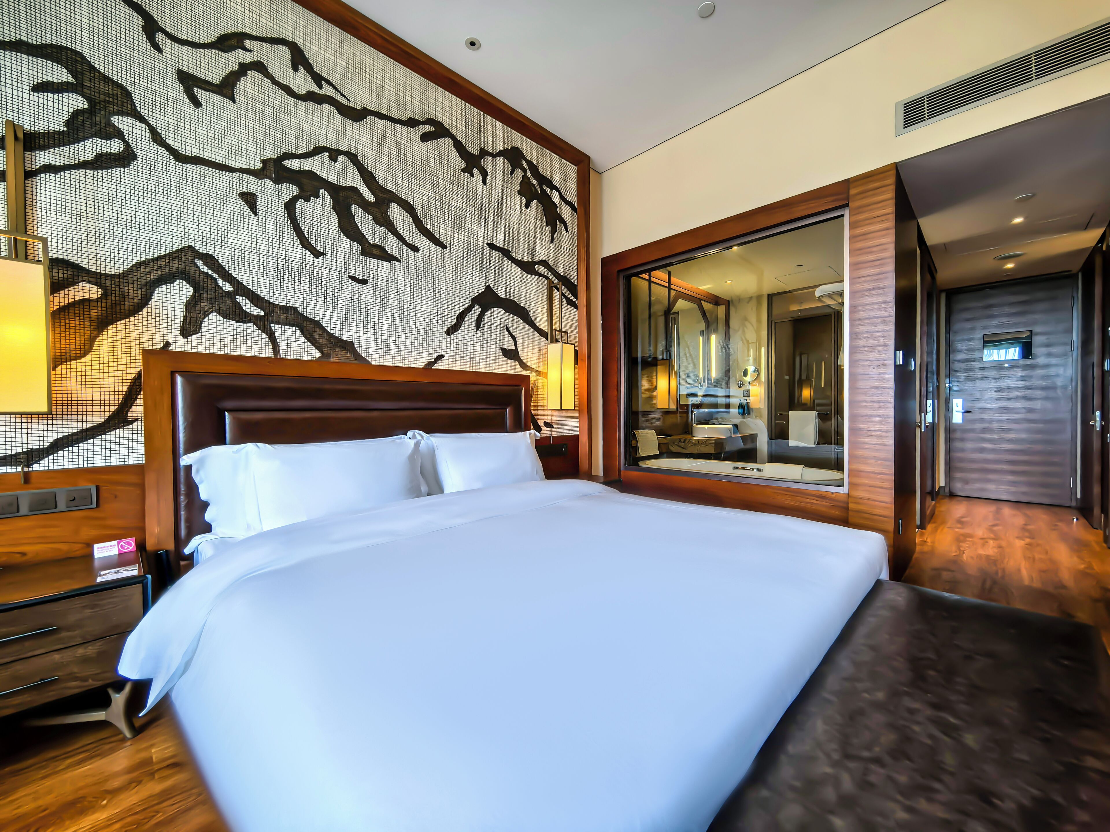 Camera d'albergo moderna con un letto grande, una testiera in legno, una parete con decoro a tema ramo d'albero, uno specchio ampio e un bagno separato visibile tramite una porta aperta.
