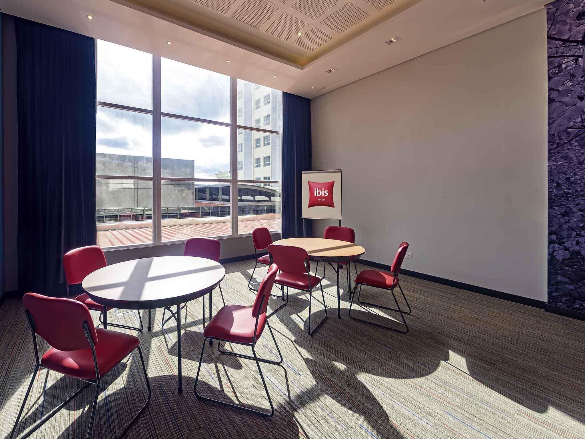 A conference room with a round table, red chairs, large windows, and a view of buildings outside.