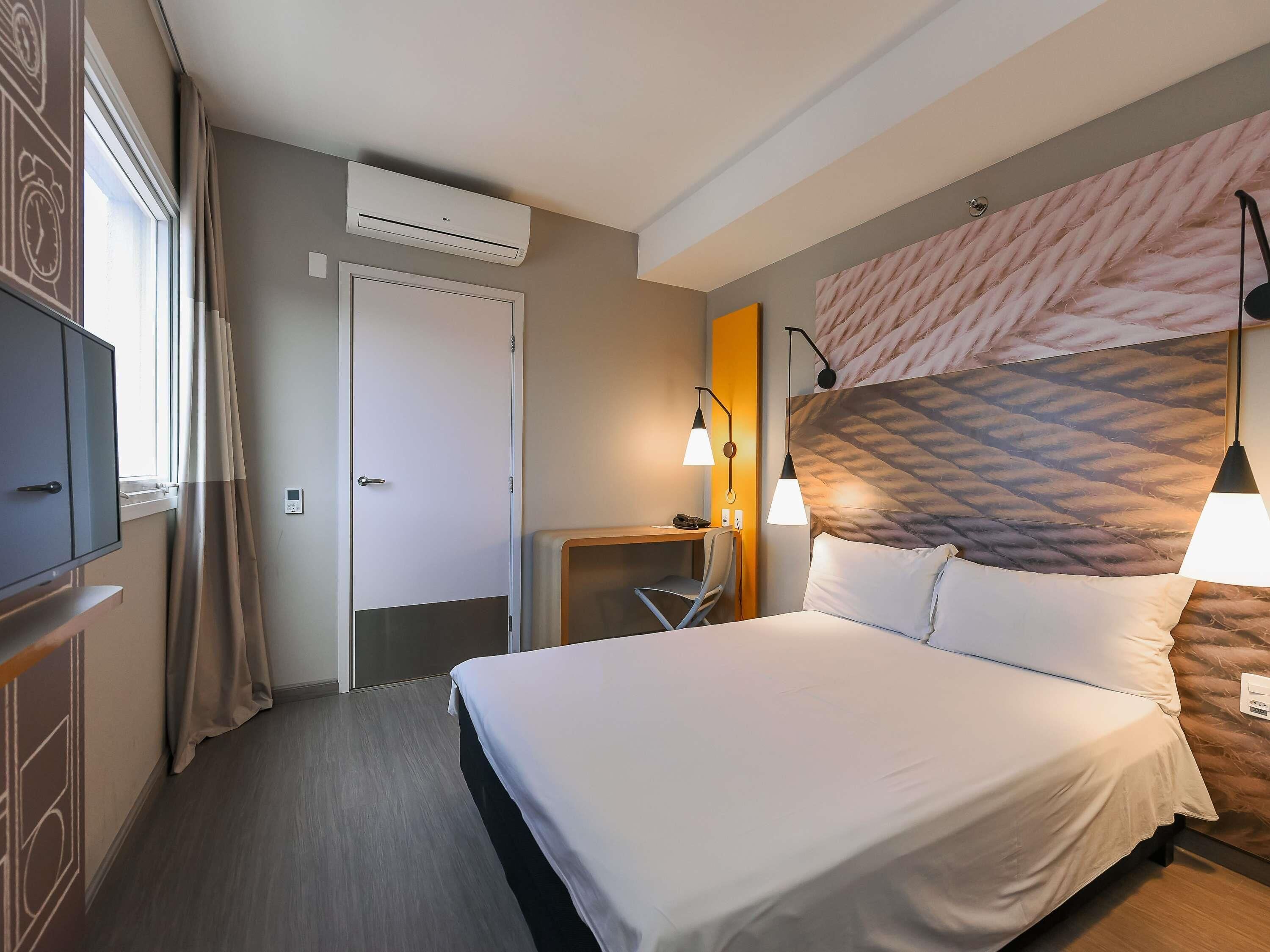 A hotel room with a bed, bedside table, lamp, air conditioning unit, and a view of the outside.