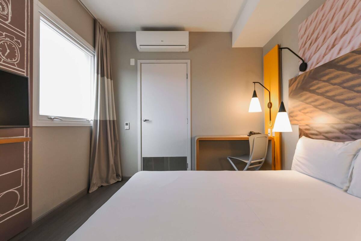 Une chambre d’hôtel comprenant un lit, un bureau avec une chaise, un appareil de climatisation et une fenêtre avec des rideaux.