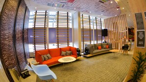 A modern lounge area with a red sofa, grey sofa, and a blue chair. There are wooden slats on the windows, a small round table, and a television on a stand. - ibis Jacarei (Jacarei)