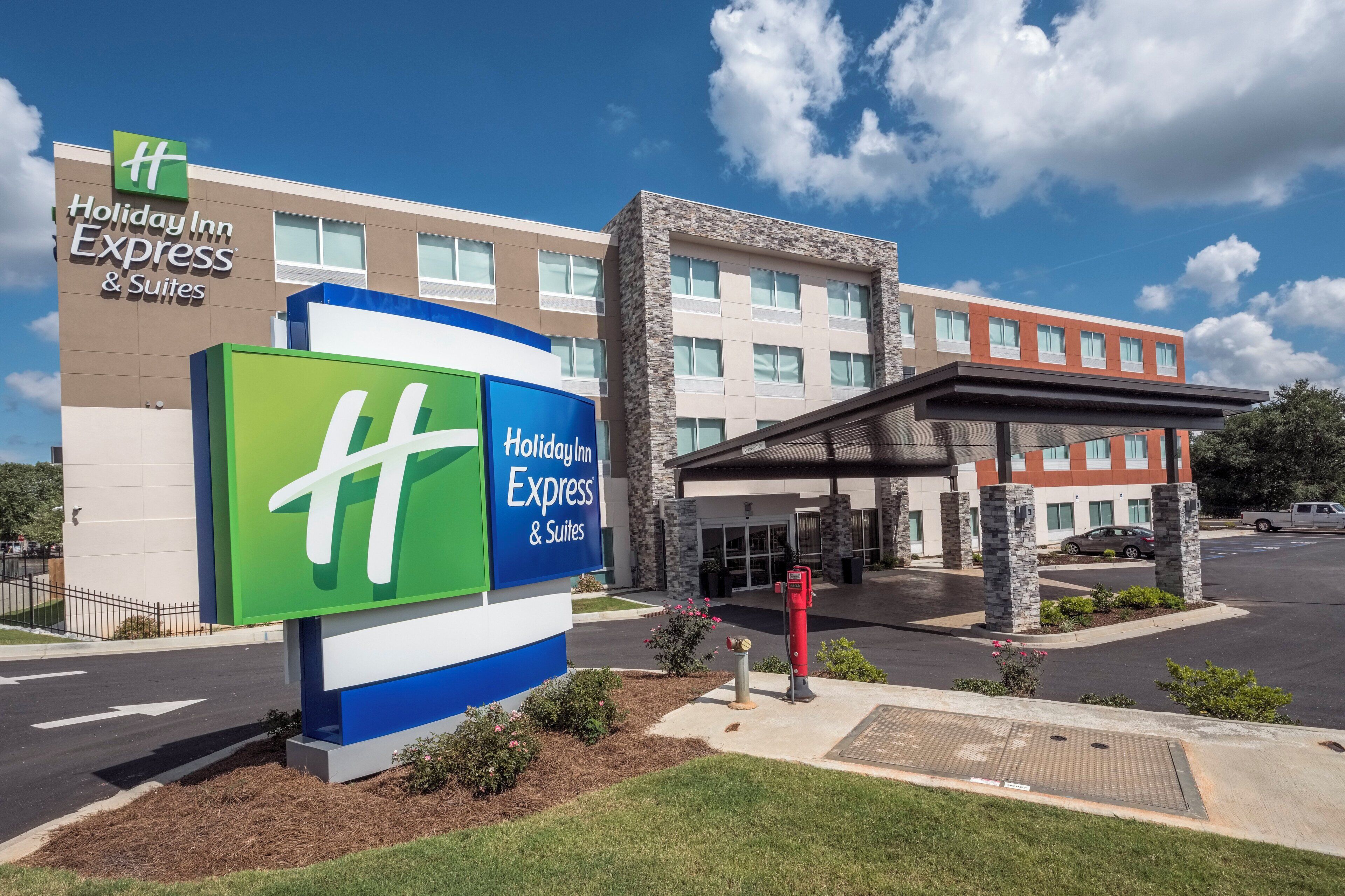 A Holiday Inn Express & Suites hotel with a prominent green and blue sign, a parking lot, and a building with a stone facade.
