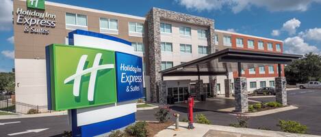 A Holiday Inn Express & Suites hotel with a prominent green and blue sign, a parking lot, and a building with a stone facade.