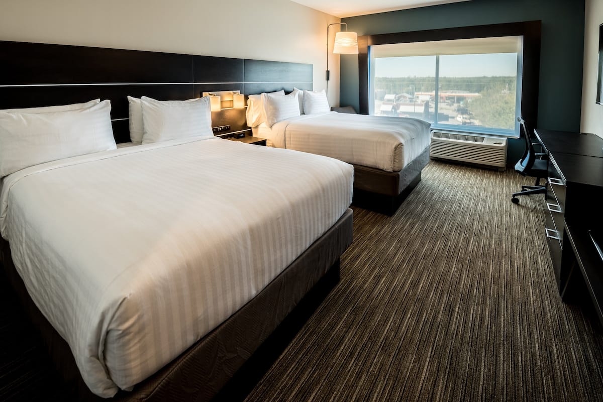 A hotel room with two beds, a desk, a chair, a large window with a view, and a wall-mounted air conditioner.