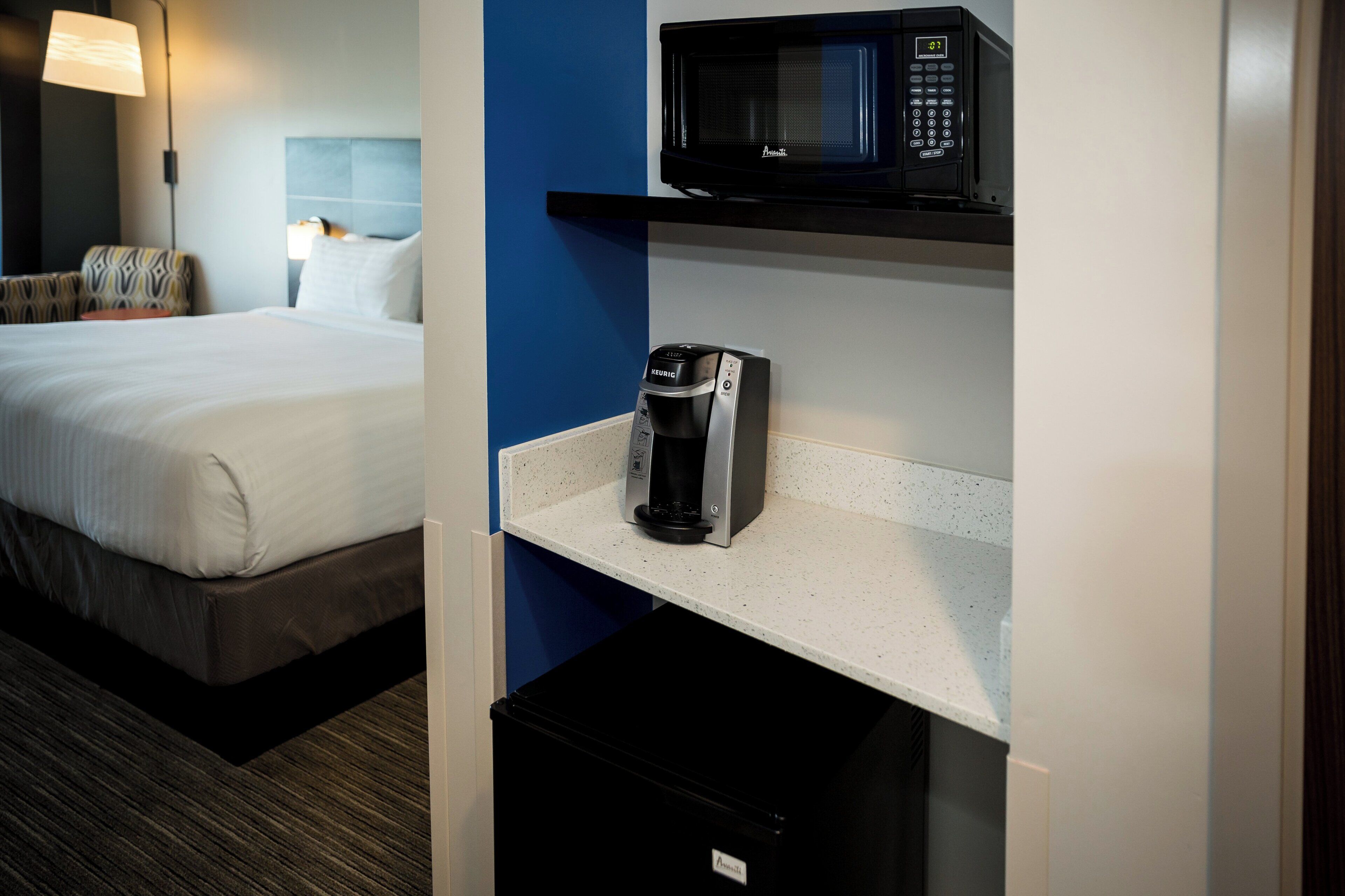 A hotel room with a bed, a minibar, a coffee maker, and a microwave on a shelf.