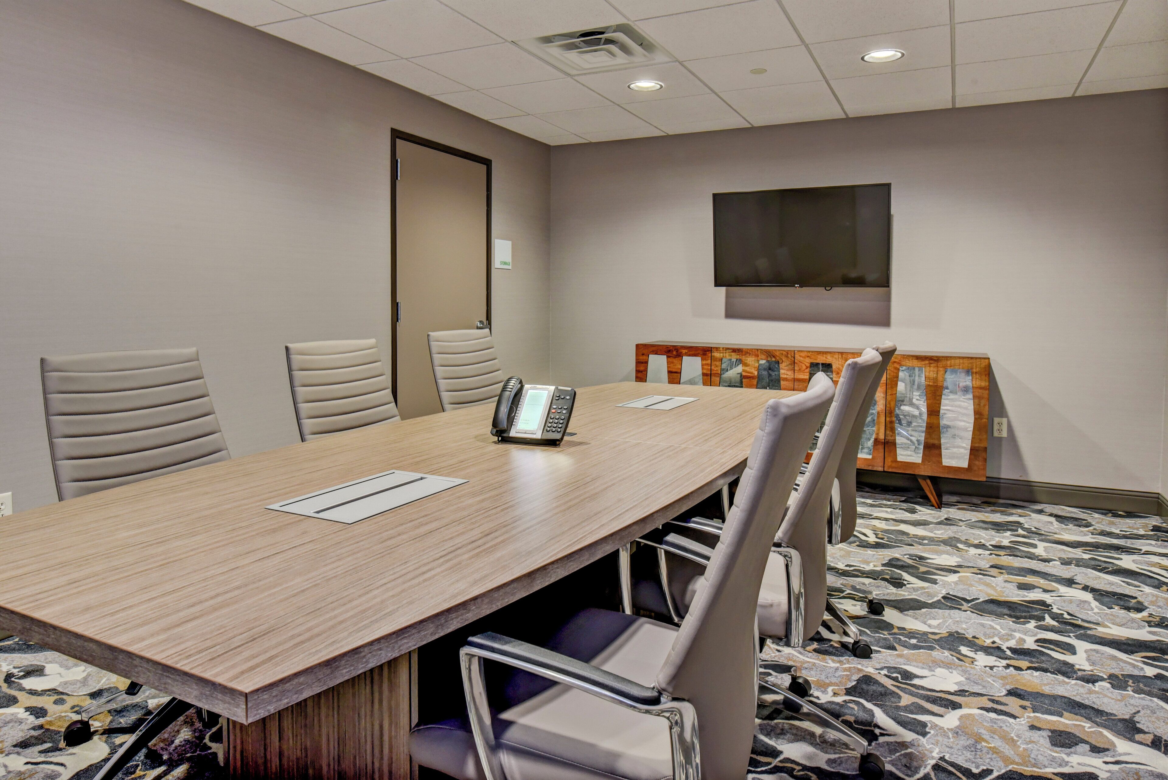 A conference room with a long table, four chairs, a phone, and a TV on the wall.