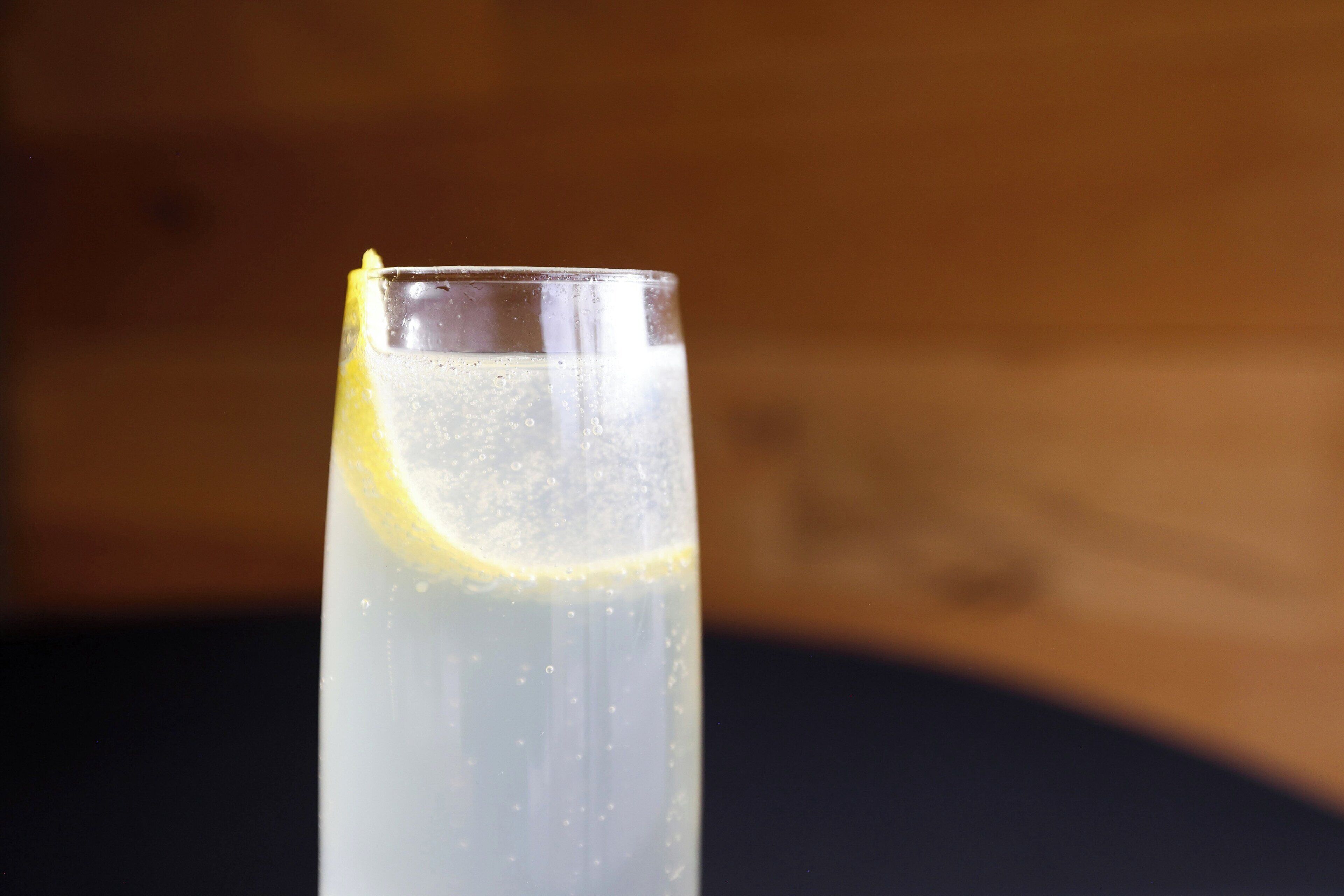 A tall glass with a lemon slice, filled with a clear liquid, possibly a beverage, against a wooden background.