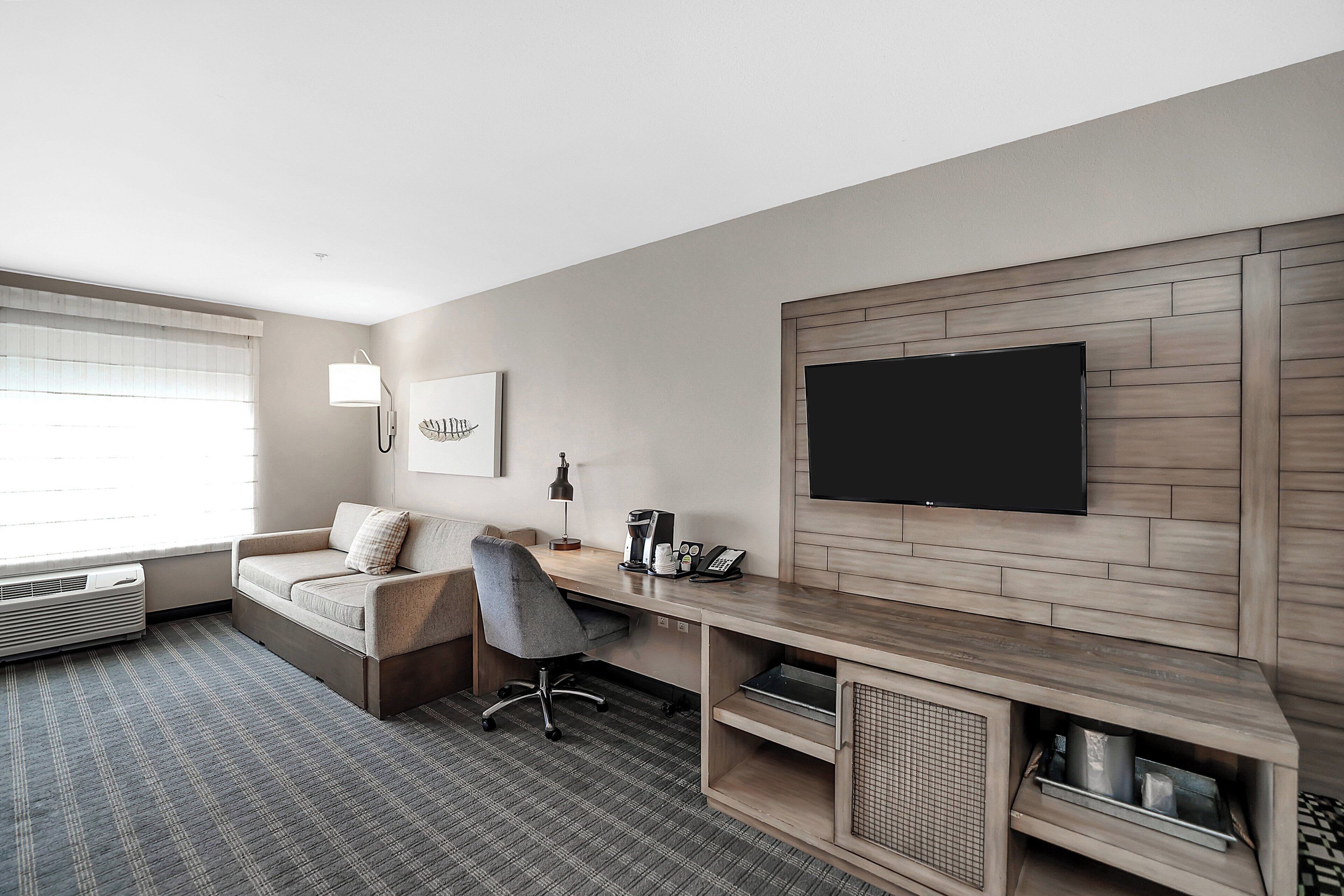 A modern hotel room with a large flat-screen TV mounted on the wall, a comfortable sofa, a desk with a lamp, and a coffee maker.